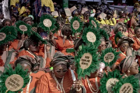 Ayo Bello / BBC A group of women in stadium seating in orange dresses holding green feather fans. 