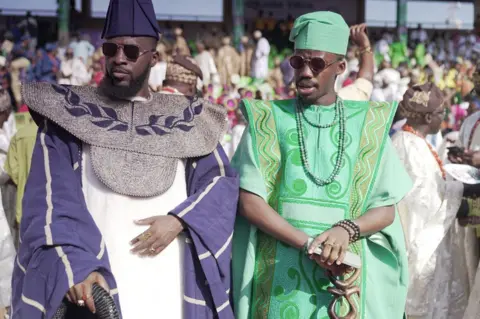 Ayo Bello / BBC Two men in traditional attire, one purple and one green, stand in the sun.