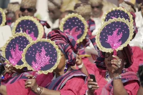 Ayo Bello / BBC Women hold up purple fans to cool themselves and shield themselves from the sun.