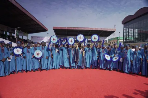 Ayo Bello / BBC A semicircle of men in blue attire holding up fans.