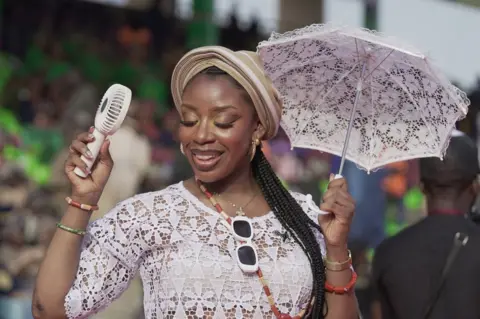 Ayo Bello / BBC A woman in white holds a small parasol and an electric fan to cool herself.